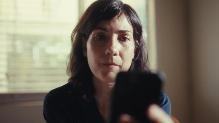 A concerned female doctor in scrubs sits at her kitchen table and reads troubling news on her smartphone, her expression growing increasingly somber