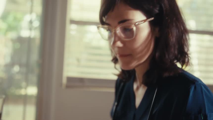 A doctor in scrubs sits at her desk calculating finances, the camera tilting up from the calculator to her focused face