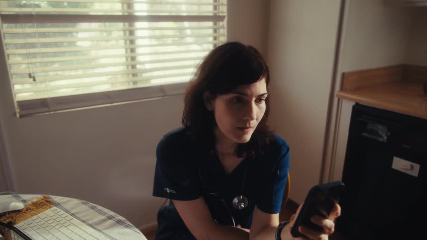 The camera slowly pushes in on a female doctor in scrubs sitting at her kitchen table looking seriously at her phone