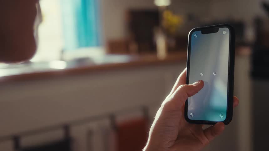 Over-the-shoulder view of a person holding a smartphone with tracking markers on the screen inside a sunlit home