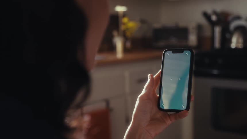 Over-the-shoulder view of a person holding a smartphone vertically, with a screen ready for compositing, inside a sunlit kitchen