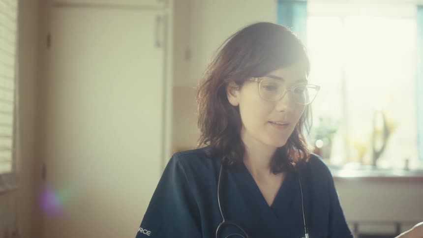 A female doctor in scrubs reviews medical documents on her tablet while working from her sunlit kitchen table at home