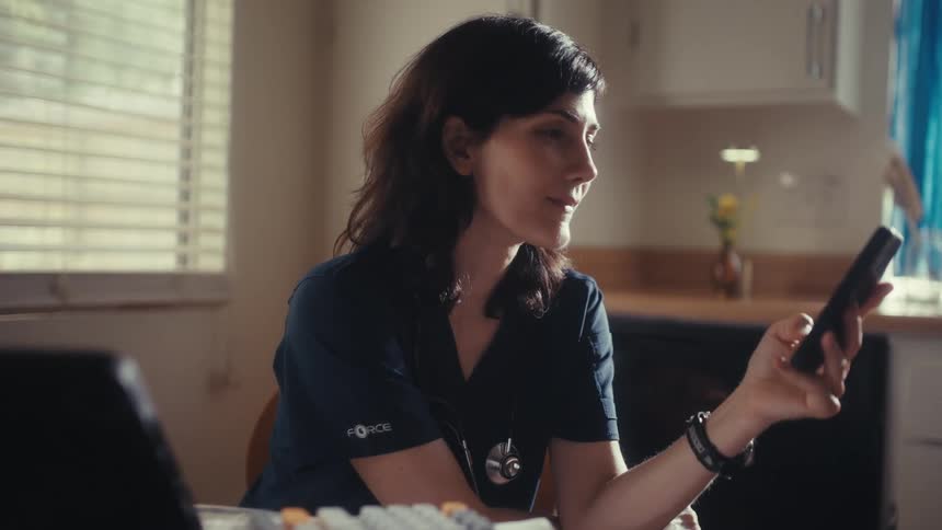 A female doctor in scrubs sits at her kitchen table and smiles warmly while looking at her smartphone