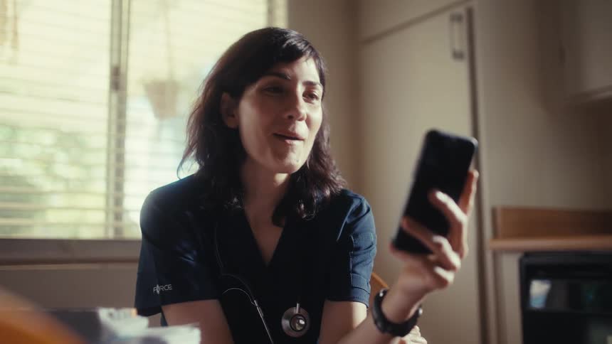 A female doctor in scrubs has a heartwarming video call on her smartphone in her kitchen, her face breaking into a wide, joyful smile