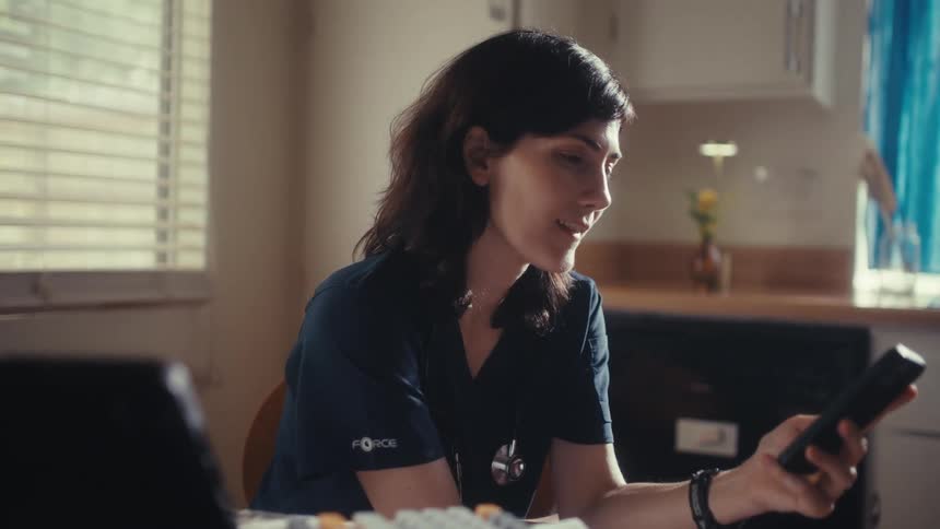 A female medical professional in scrubs works remotely from her sunlit kitchen, smiling as she uses her smartphone