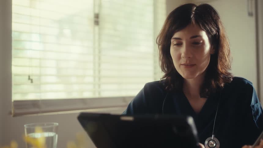 A smiling female doctor in scrubs uses a digital tablet while sitting at her desk in a sunlit office