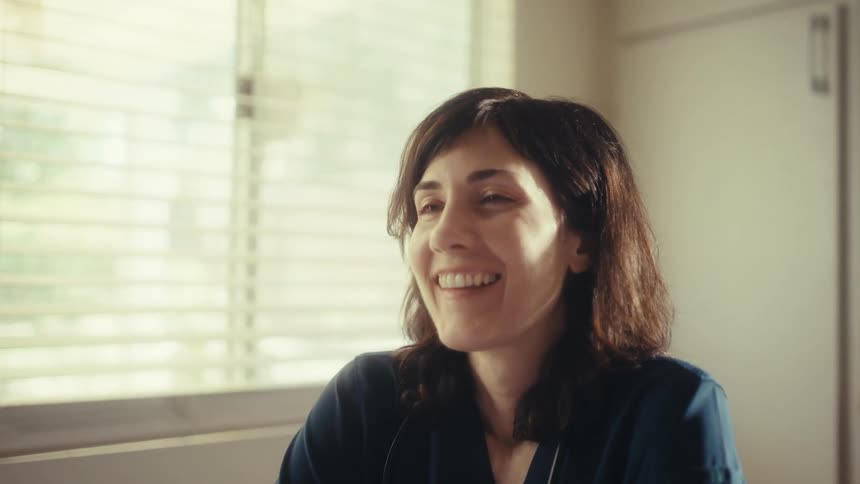 A female doctor in scrubs smiles while using a tablet at her sunlit kitchen table, with the camera slowly zooming into a close-up