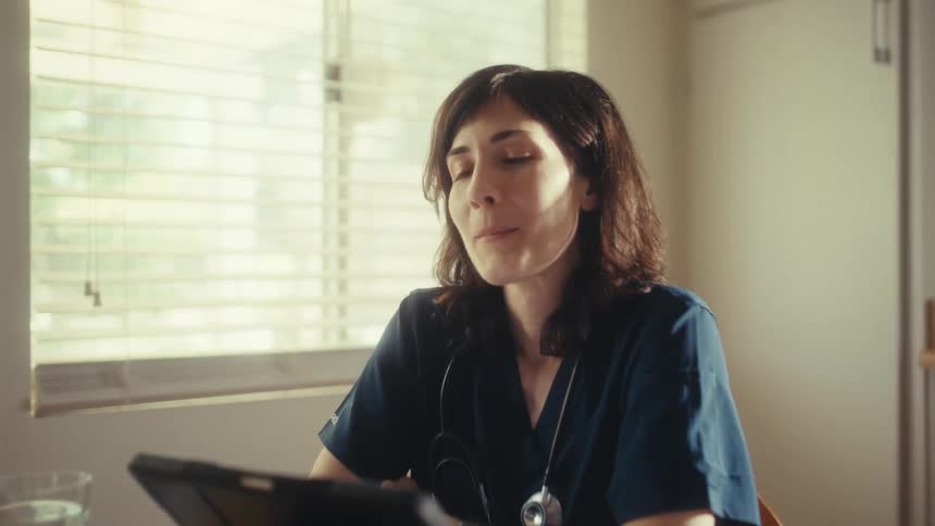 A smiling female doctor in scrubs uses a tablet while working from her sunlit kitchen table, feeling content and relaxed