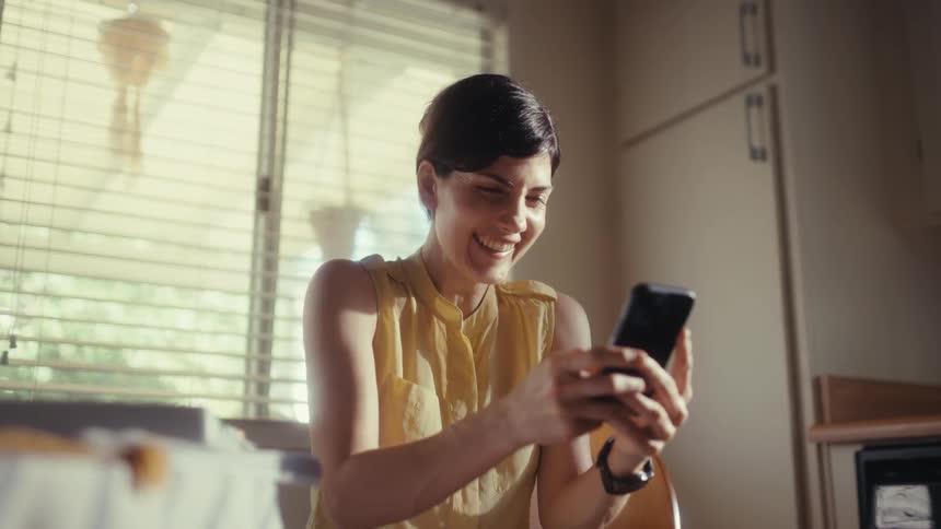 A happy woman with short hair smiles and laughs while looking at her smartphone in a sunlit room