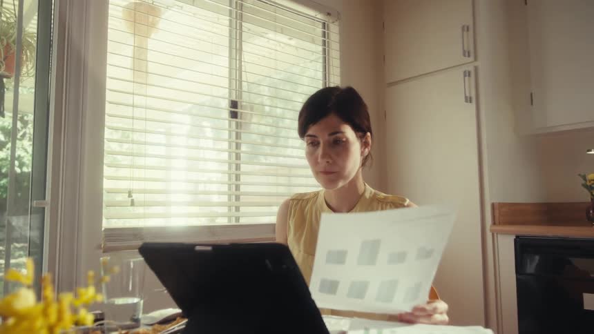 A focused woman analyzes charts and graphs on documents while working from her sunlit kitchen table at home