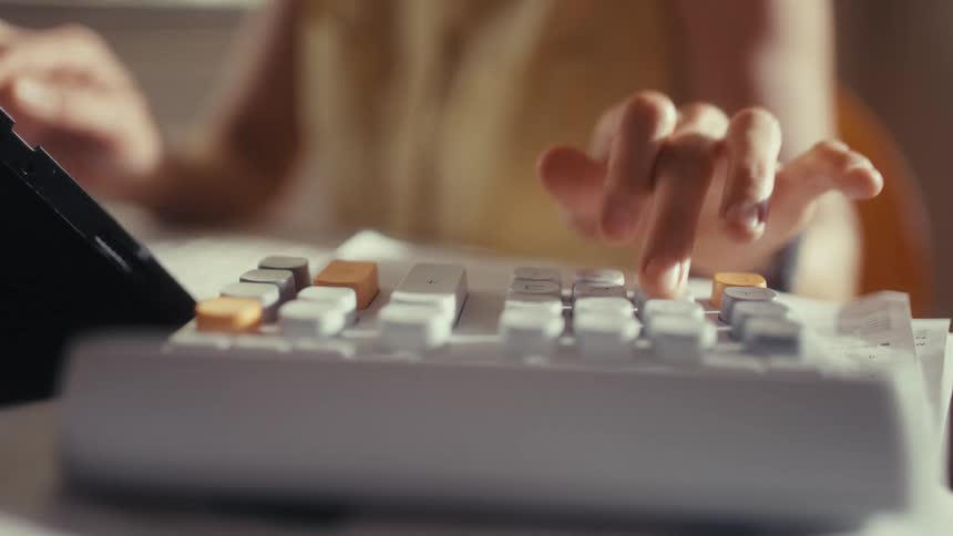 A close-up shot of a woman's hands using a white calculator at a desk in slow motion