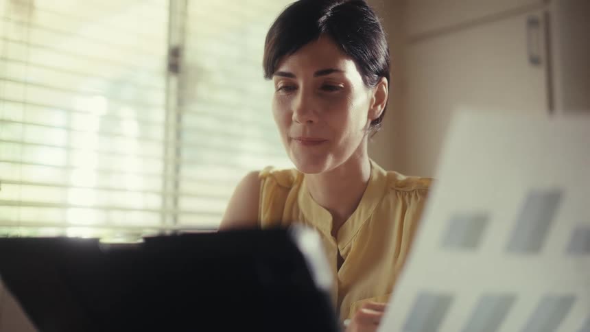 A businesswoman with short dark hair smiles while working on her tablet in her sunlit office