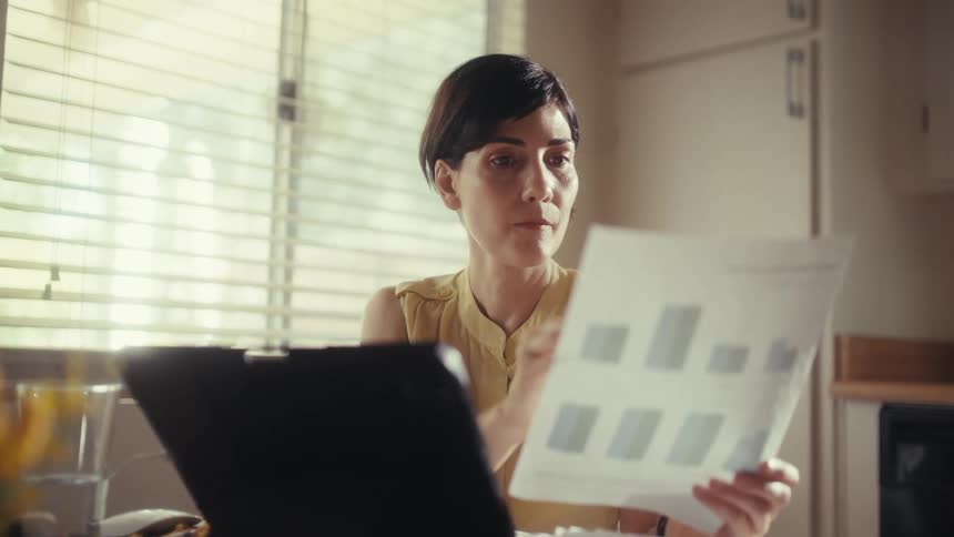 A focused woman analyzes financial documents and bar charts while working from her sunlit kitchen table