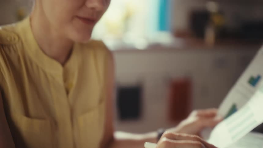 A focused businesswoman analyzes financial charts on paper while working on her tablet in a brightly lit home office