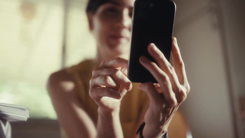 A smiling woman scrolls on her smartphone in a sunlit room, with the focus shifting to her hands