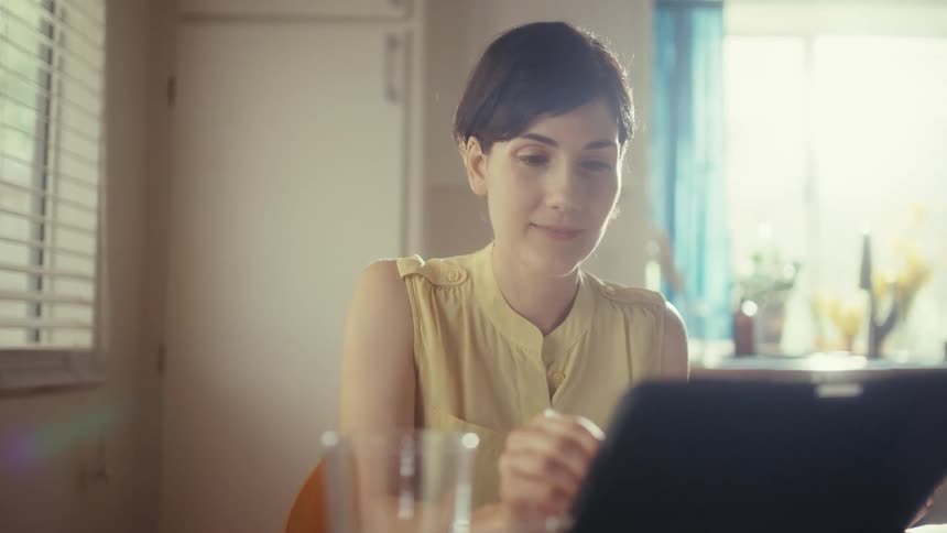 A focused woman works from home on her tablet with a stylus, sitting at a sunlit dining table in her kitchen