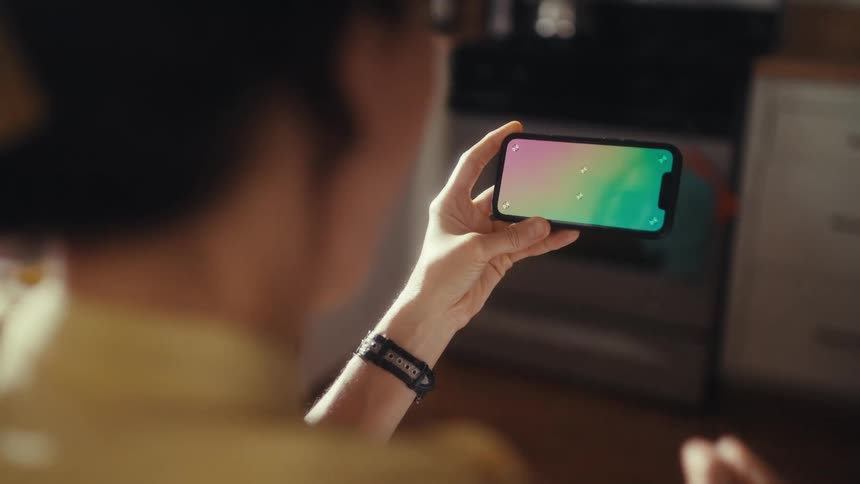 Over-the-shoulder shot of a person celebrating while watching content on a smartphone with a green screen in their kitchen