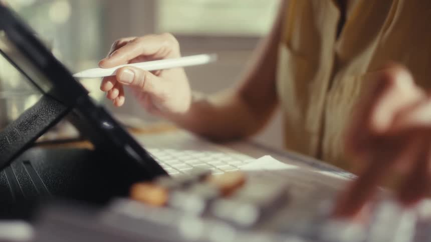 Close up of a woman multitasking at her desk, using a stylus on a tablet while calculating finances on a calculator