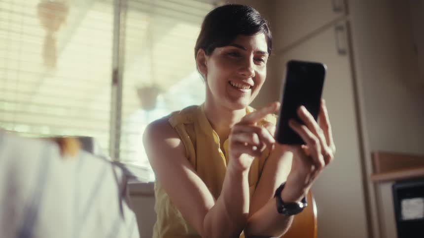 A smiling woman in a yellow top enjoys scrolling on her smartphone in a sunlit room