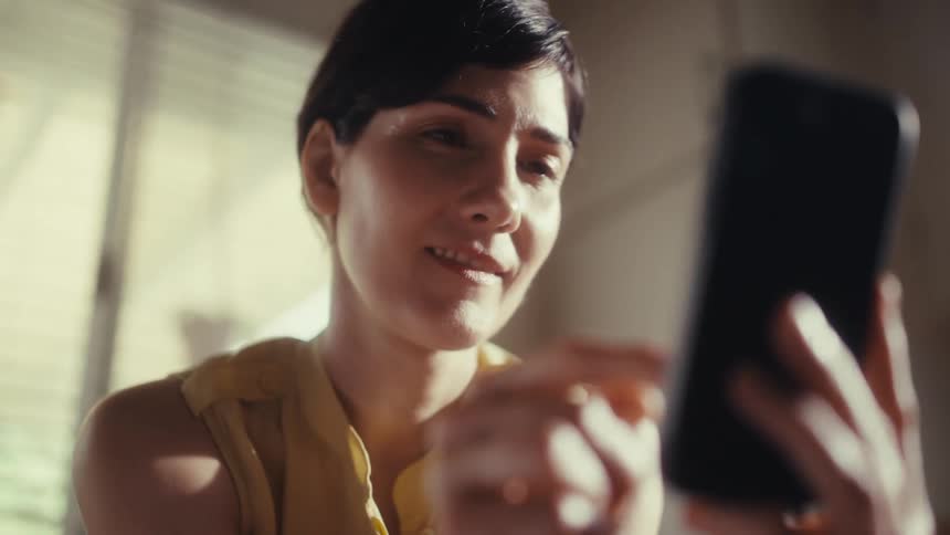 A woman with short dark hair smiles while scrolling on her smartphone in a sunlit room