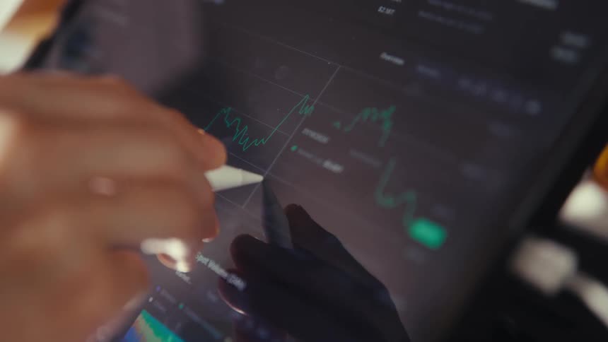 A close-up view of a person's hand using a stylus to analyze fluctuating financial market charts on a tablet screen