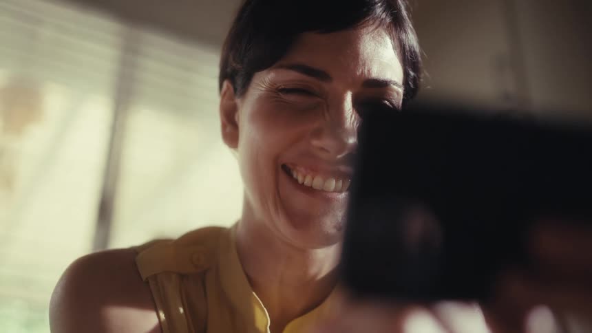 An attractive woman with short dark hair smiles and laughs with joy while watching a video on her smartphone