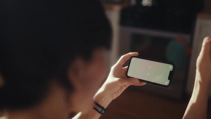 Over-the-shoulder view of a woman holding a smartphone with a green screen, grooving to the rhythm in a sunlit kitchen
