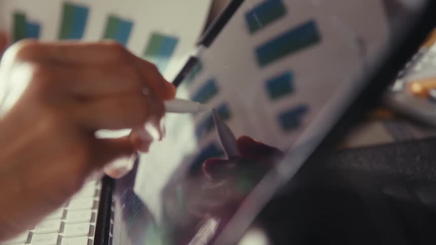 A person analyzes financial charts on a tablet with a stylus while cross-referencing a printed report