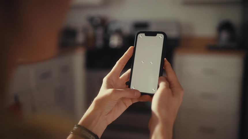 A close-up over-the-shoulder shot of a person holding a smartphone with a blank screen and tracking markers, tapping the screen in their home