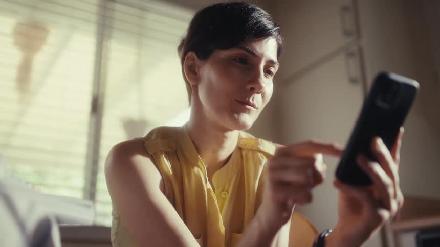A woman with short dark hair smiles while scrolling on her smartphone, enjoying content in a room bathed in warm, natural light