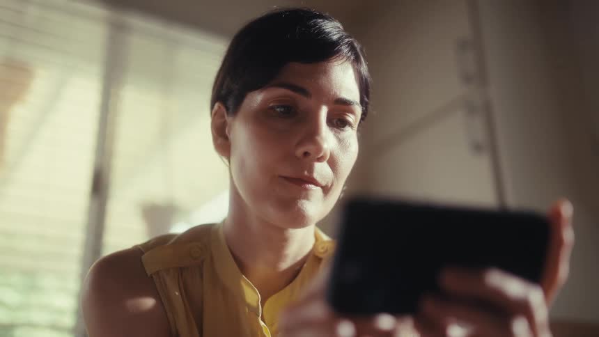 A woman with short dark hair intently watches content on her smartphone, illuminated by soft window light