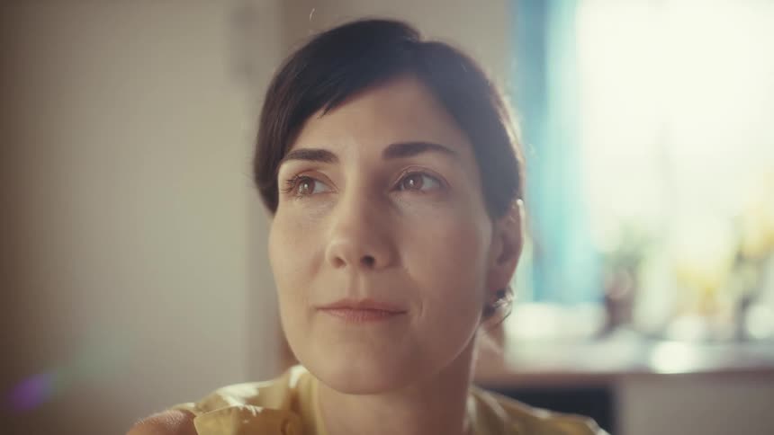 A serene woman with a pixie cut sits in a warmly lit room, her face full of quiet, wistful contemplation