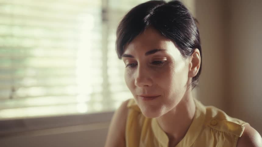 A focused woman works from home at her sunlit dining table, analyzing charts with a contented smile as the camera slowly zooms in