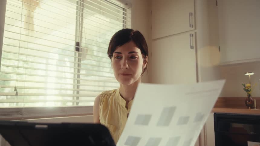 A focused woman works from home at her sunlit kitchen table, analyzing charts on paper and her laptop