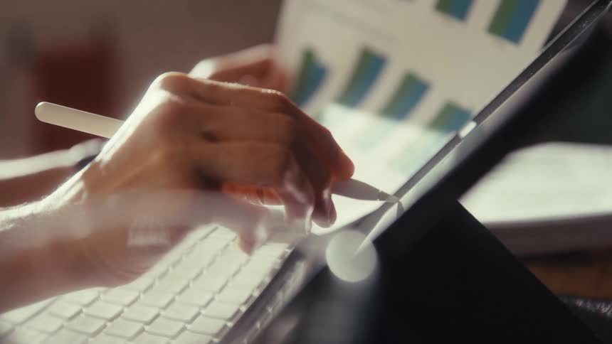 A person analyzes charts and graphs on a tablet using a stylus while working at a sunlit desk