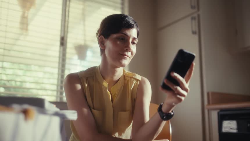 A short-haired woman in a yellow top sits in her sunlit kitchen, smiling as she scrolls through her smartphone