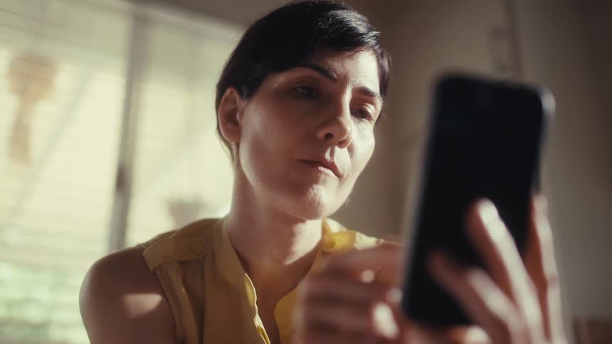 A woman with a focused expression scrolls through her smartphone in a sunlit room