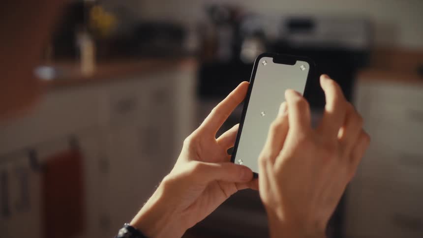 An over-the-shoulder shot of a person using a smartphone with a blank screen and tracking markers in a kitchen