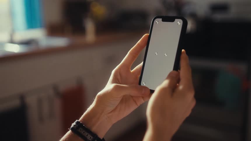 Over-the-shoulder view of a person's hands holding a smartphone with a blank screen and tracking markers in a sunlit room