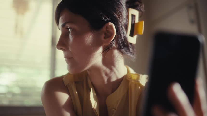 A woman with a serious and contemplative expression looks at her smartphone screen in a sunlit room