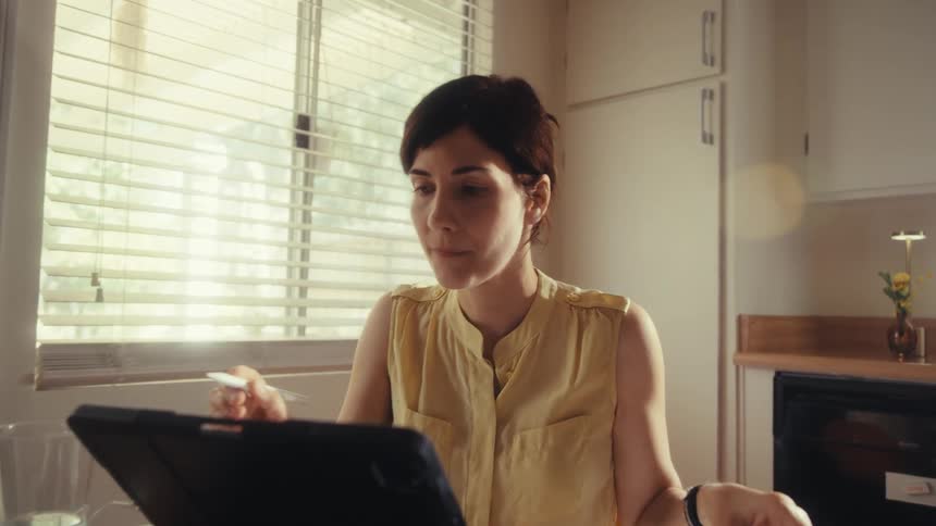 A focused woman works from home at her sunlit kitchen table, using a tablet with a stylus while analyzing charts