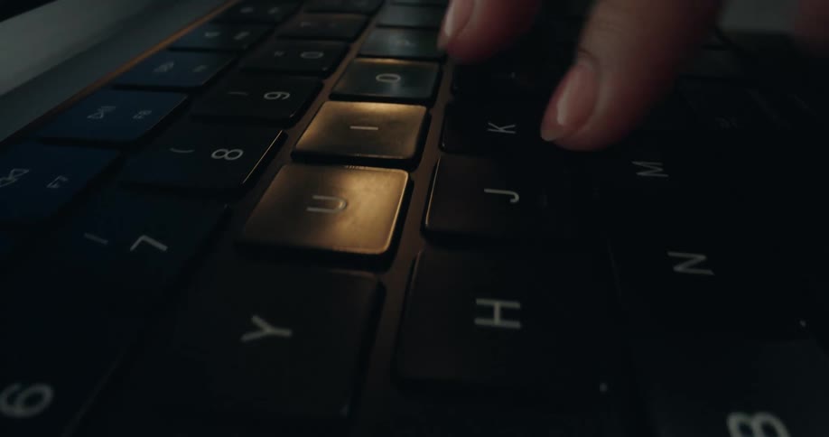 A dramatic close-up shot of fingers typing on a black laptop keyboard in a dimly lit room