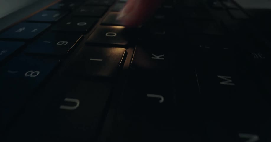 A macro pan shot captures fingers typing on a black laptop keyboard in a dimly lit environment