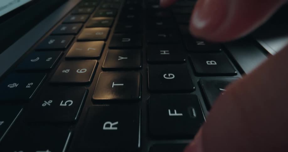 Extreme close-up dolly shot of a person's manicured fingers typing on a laptop keyboard in a dark room