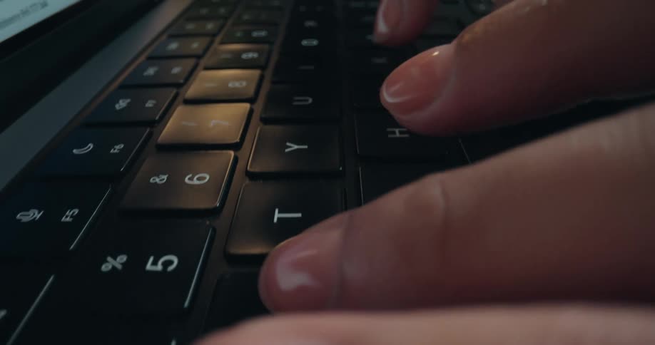 Fingers with clear nail polish type on a black laptop keyboard in a slow-motion, low-angle macro shot
