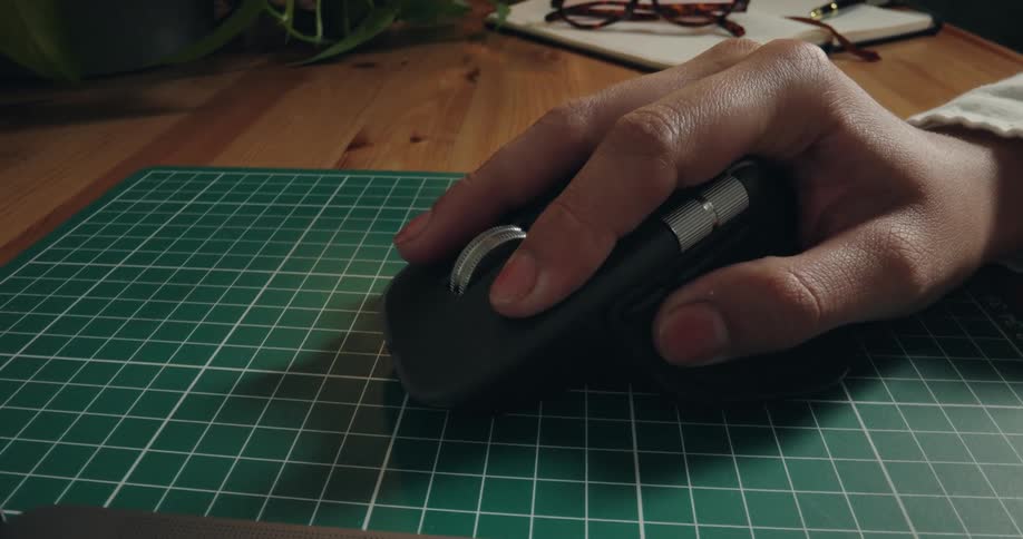 Close-up of a person's hand navigating with an ergonomic wireless mouse on a green cutting mat while working at their desk