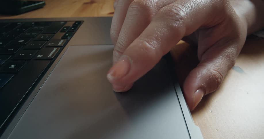 A close-up panning shot of a person's hand using the trackpad on a modern laptop resting on a wooden desk