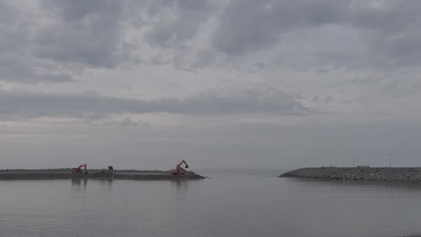 Excavators work on constructing a stone breakwater at a harbor entrance under a moody, overcast sky
