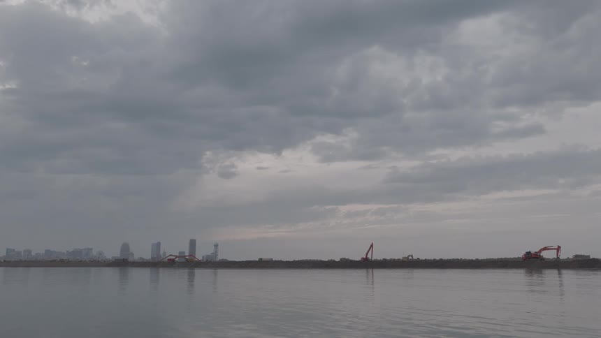 A distant city skyline is visible across the water behind a construction site with excavators under a dark, cloudy sky