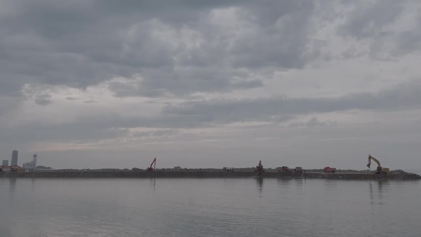 Heavy machinery works on a coastal land reclamation project on a peninsula under a grey, overcast sky